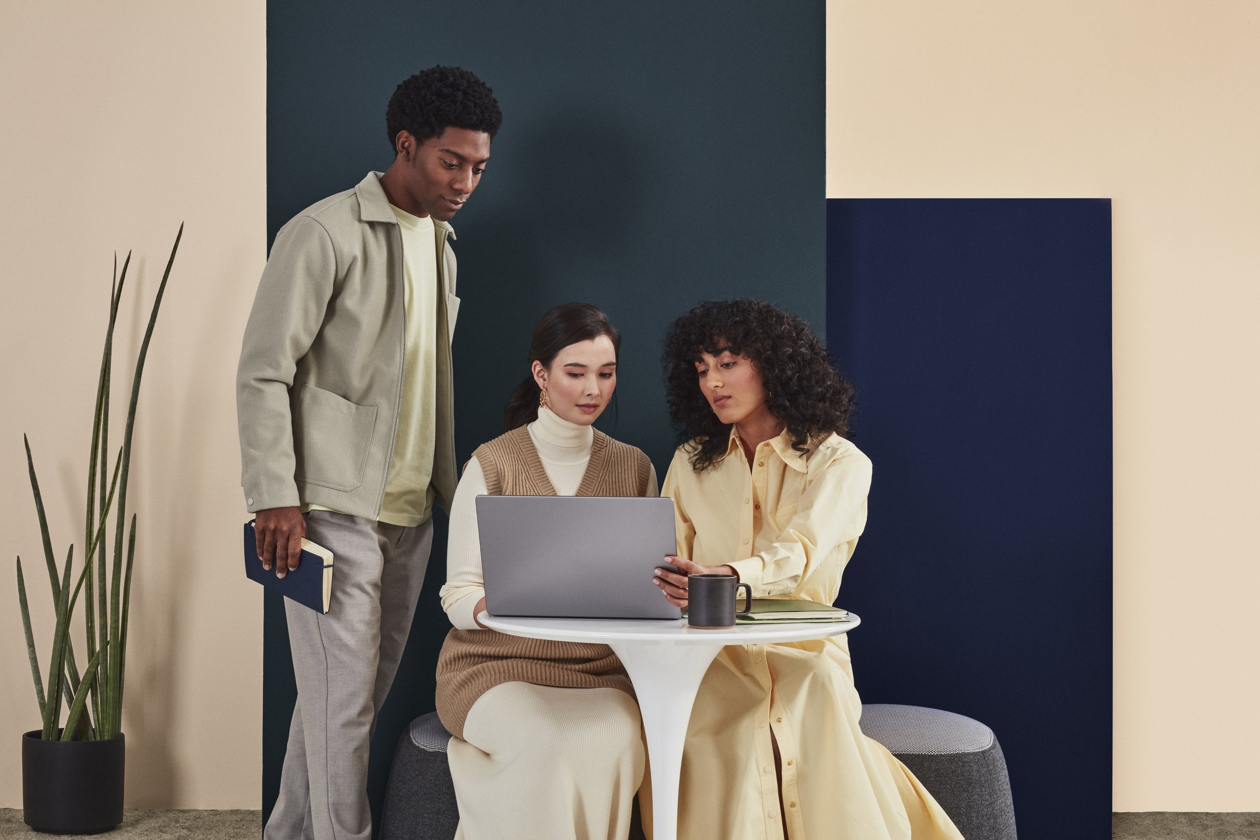 Three people gathered around a laptop at a small round table in an office space with colorful walls and a tall potted plant in the background.