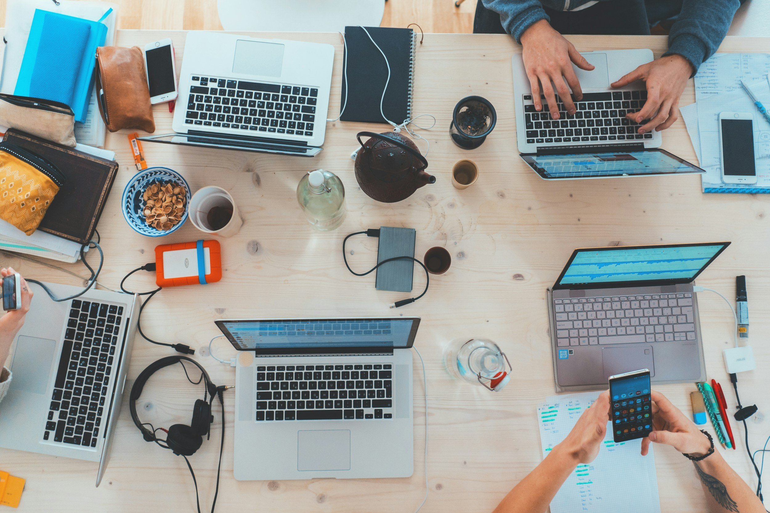 A top-down view of a wooden table with multiple open laptops, smartphones, notebooks, pens, and drinks, showing a busy workspace with people working on laptops and using phones.