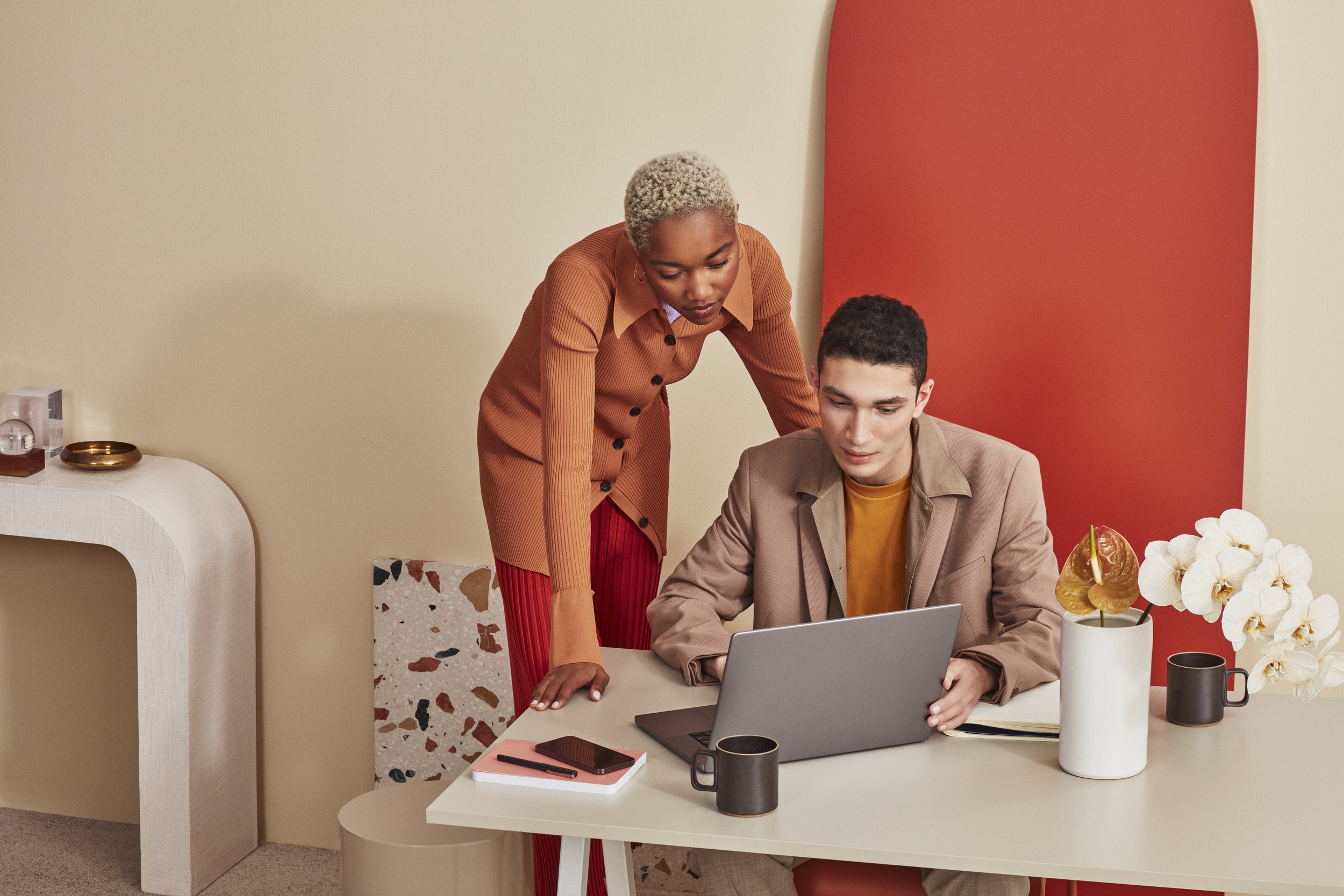 A woman stands beside a man who is sitting at a table, both looking at a laptop, with a notebook, two mugs, and a potted plant on the table.